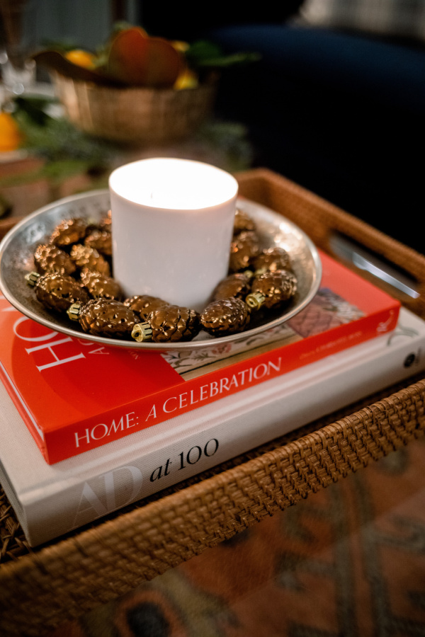 Candle in silver dish with ornament pinecones on books in tray.