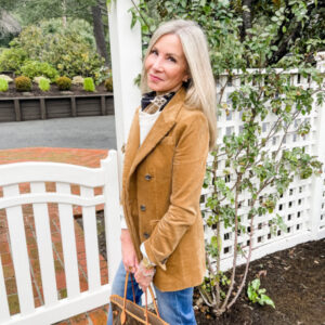 woman wearing corduroy blazer and jeans standing in front of white fence.
