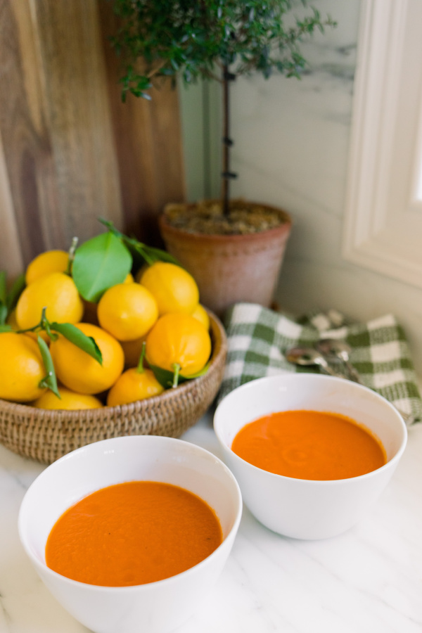 Two bowls of tomato soup sitting next to a bowl of lemons and a topiary.
