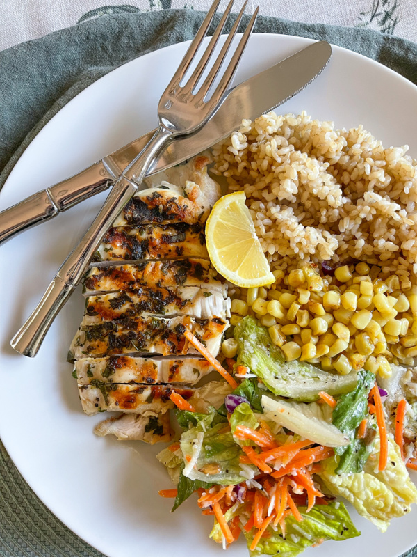 Grilled herbed chicken on plate with rice, corn, salad and lemon wedge.