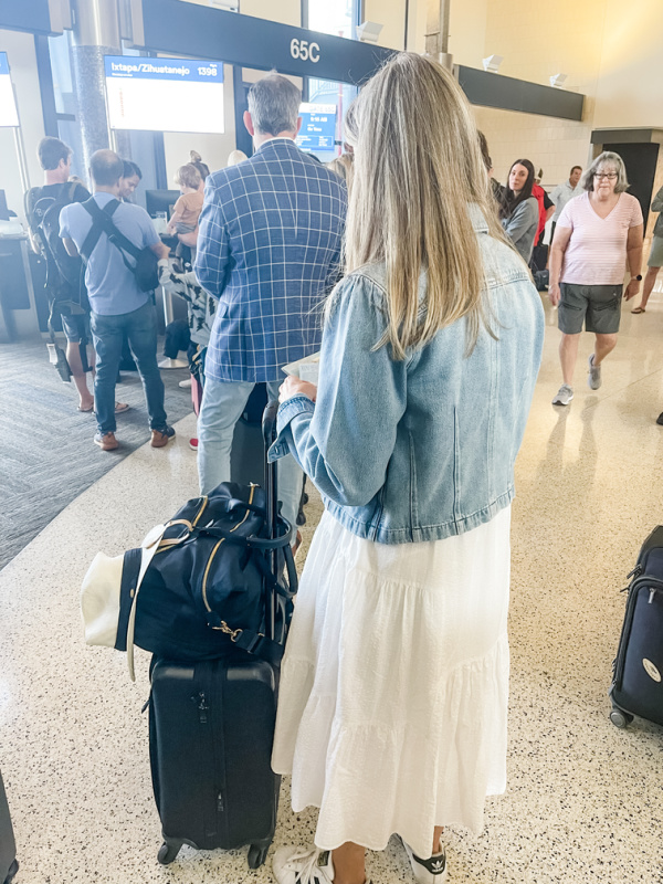 Woman wearing white dress and denim jacket standing with luggage waiting to board plane.