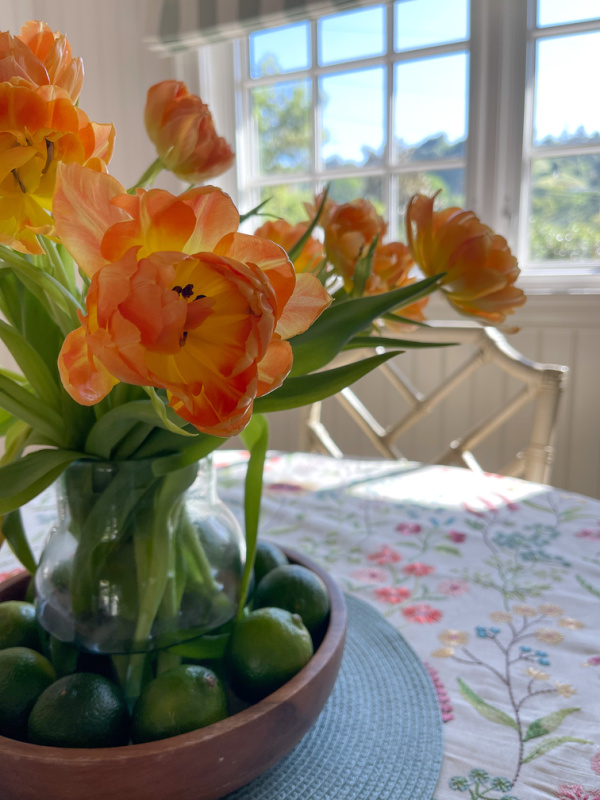 Vase of orange tulips surrounded by limes on an embroidered table cloth.