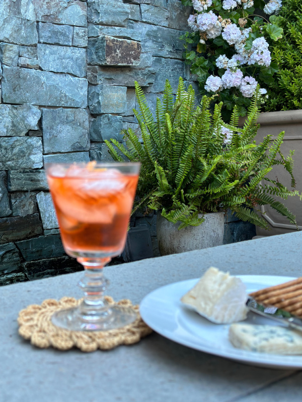 Aperol Spritz and cheese plate on backyard table in front of fountain.