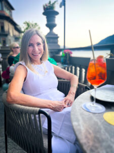 Woman wearing white dress sitting at table overlooking Lake Como with an Aperol Spritz.