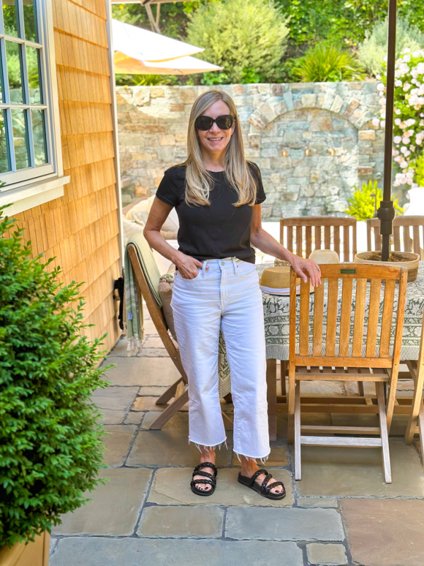 Woman wearing black t-shirt and white jeans standing next to table in backyard.