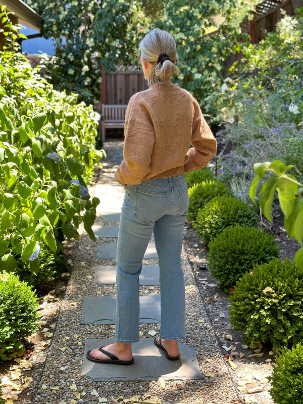 Woman standing in garden with back to camera wearing jeans, flip flops and a fall cardigan.