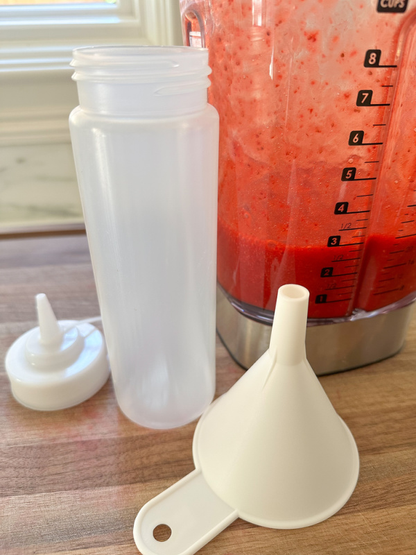 Blender full of strawberry puree sitting next to squeeze bottle and funnel.