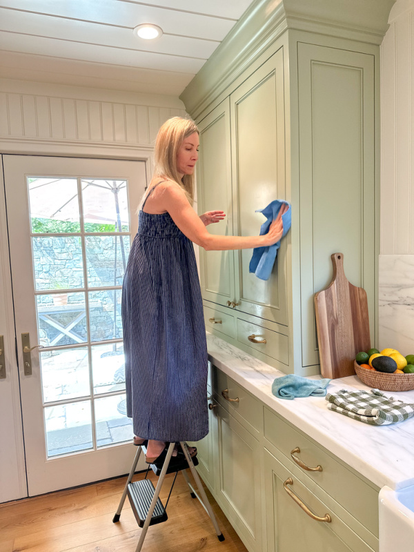 Woman on step stool in kitchen washing cabinetry.