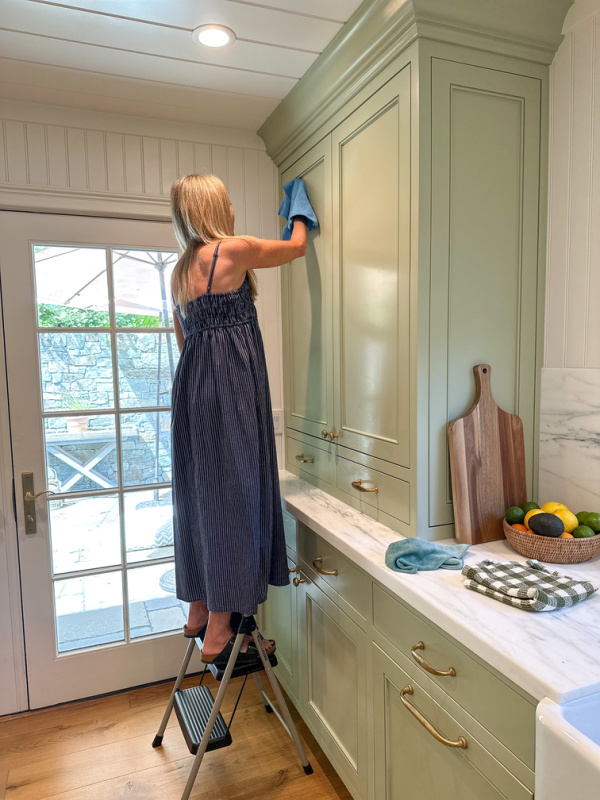 Woman on step stool cleaning kitchen cabinet fronts.