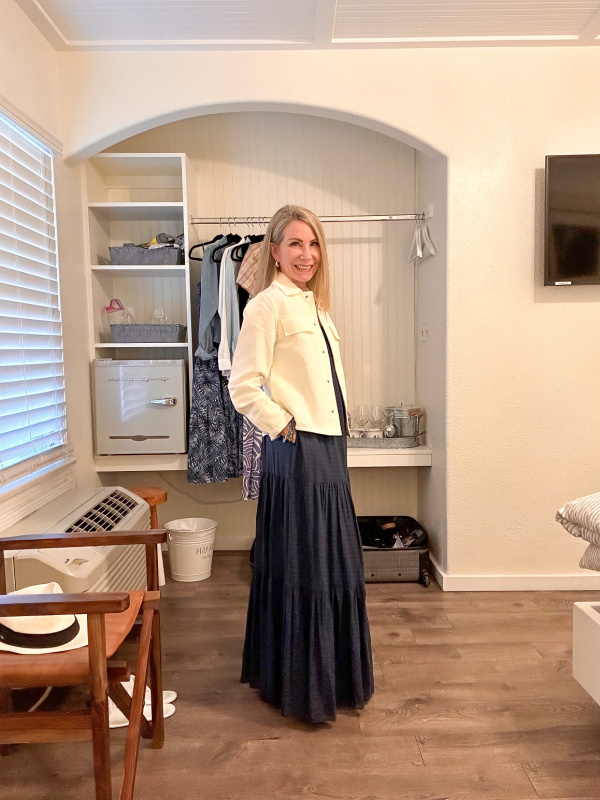 Woman wearing blue maxi dress and ivory jacket in hotel room.