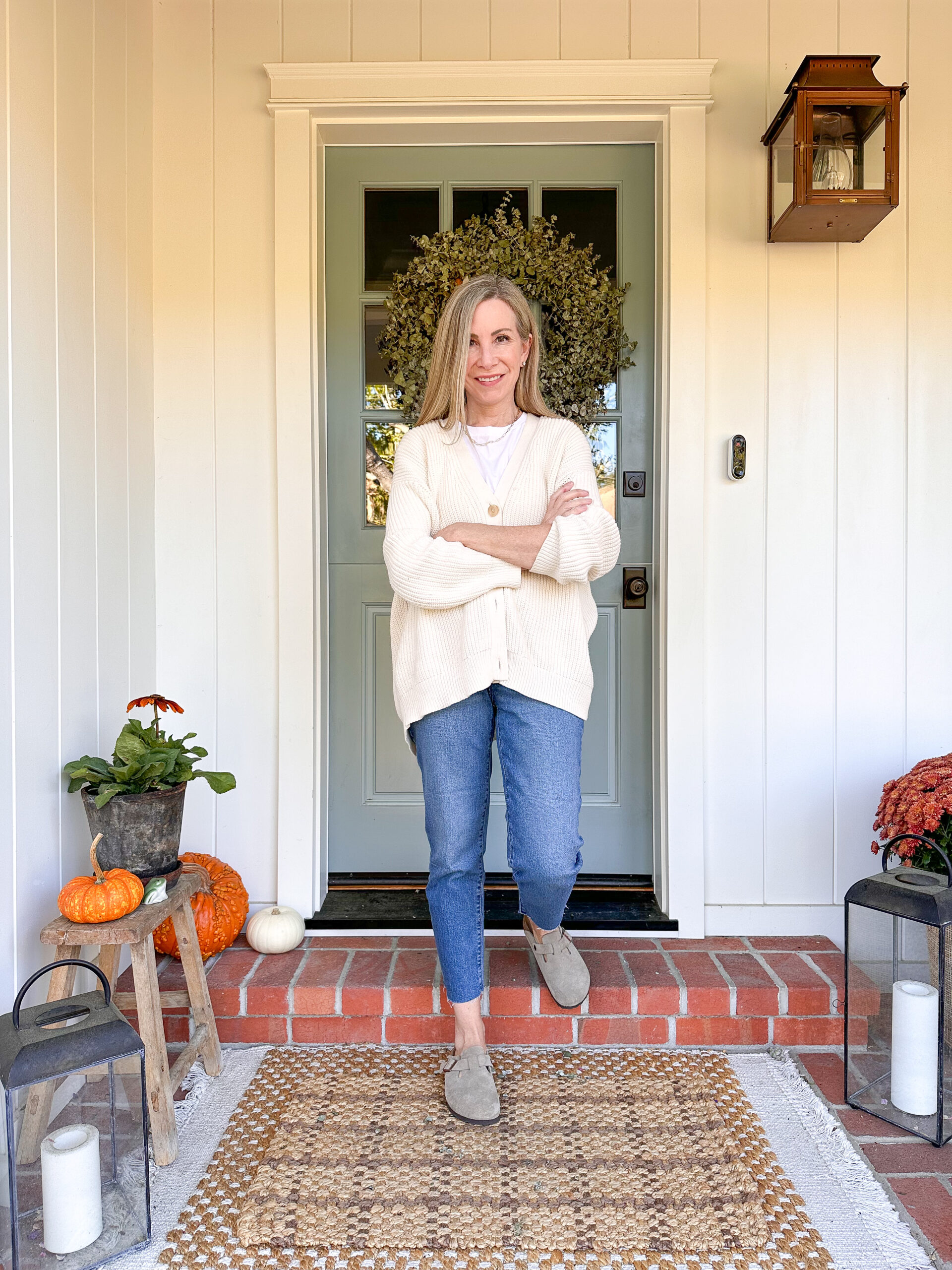 Woman standing on porch in front of blue Dutch door surrounded by fall decor.