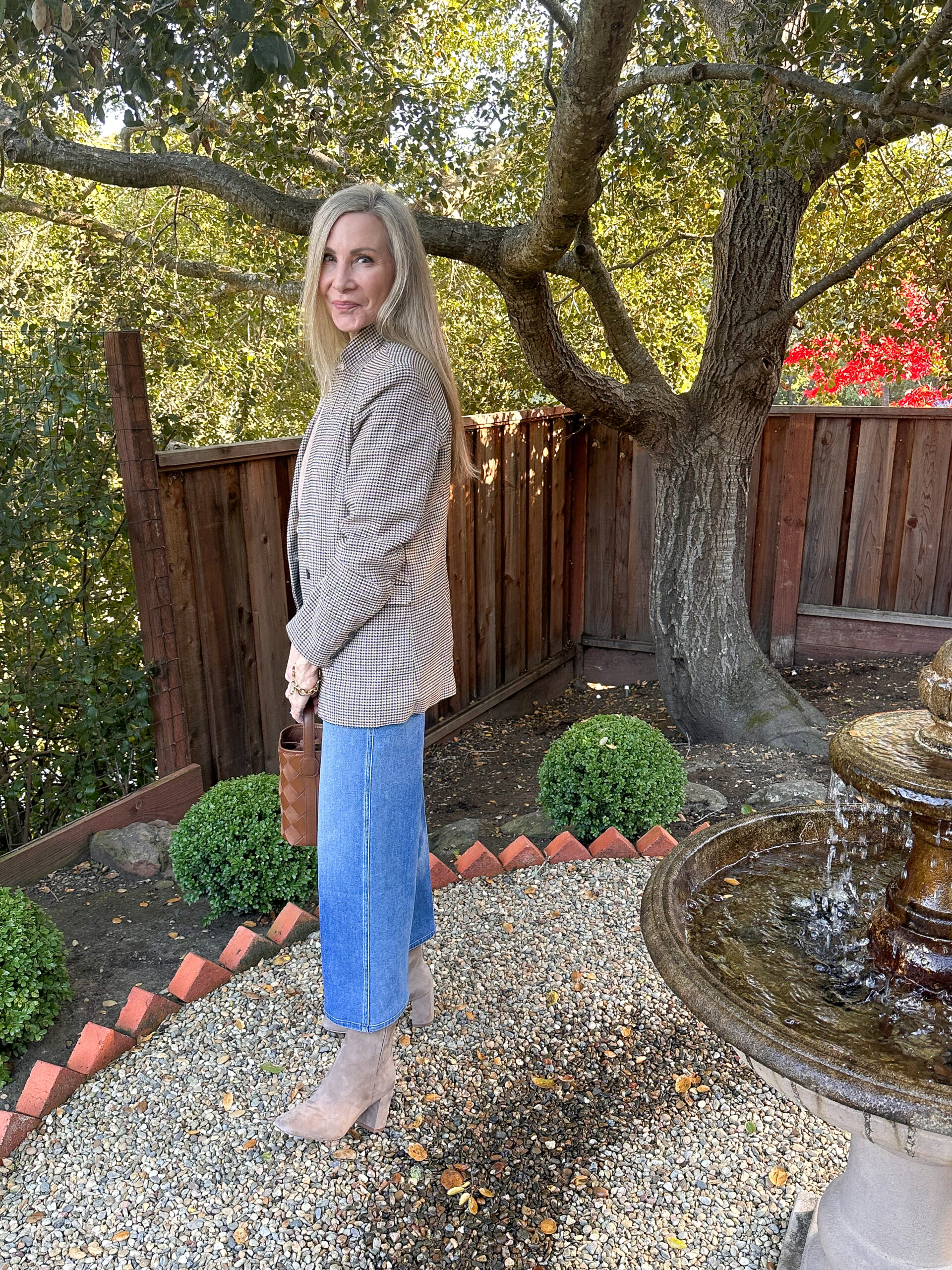 Woman standing in gravel garden path wearing jeans and a blazer.