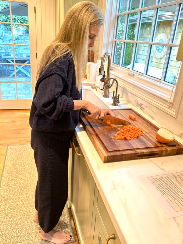 Woman standing in kitchen chopping carrots on cutting board.