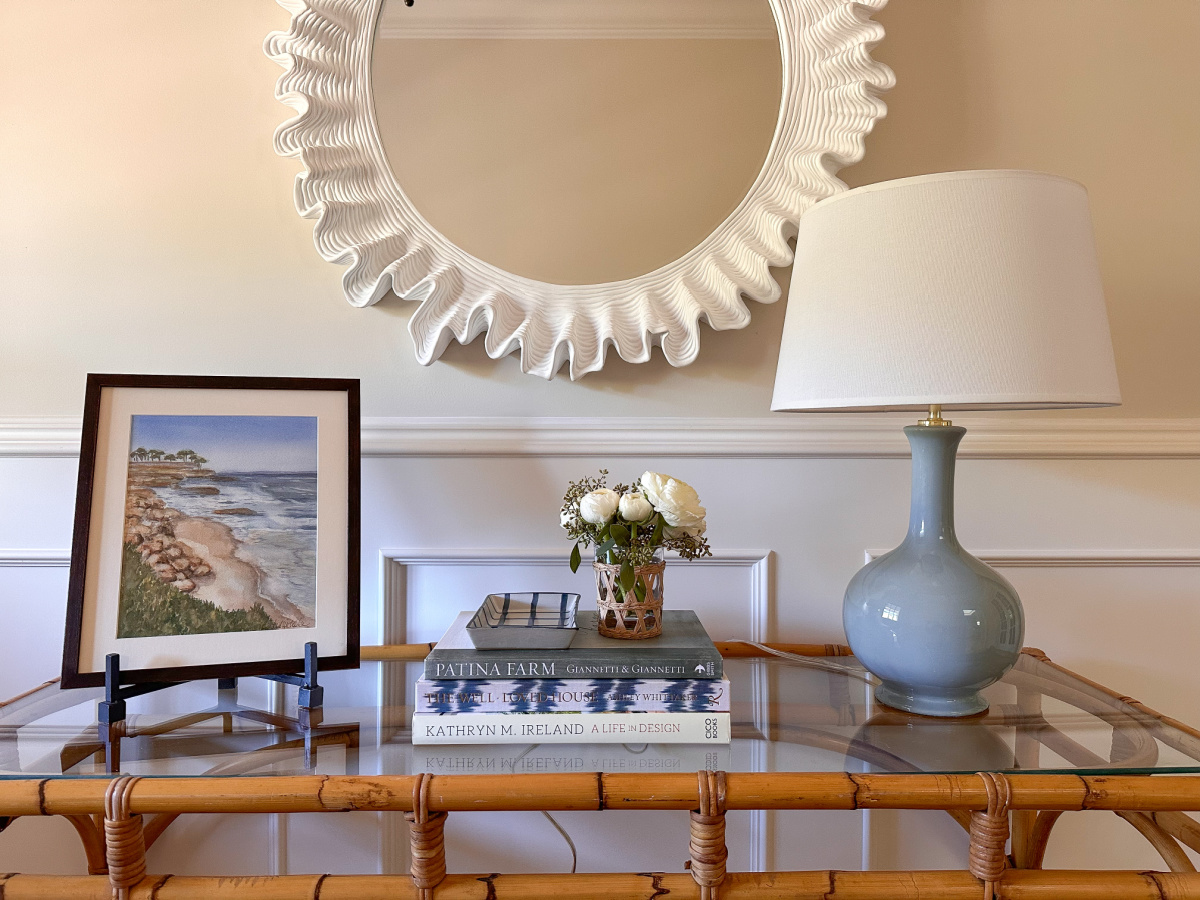 Bamboo entryway table styled with art, books and a lamp, and scalloped mirror on wall.