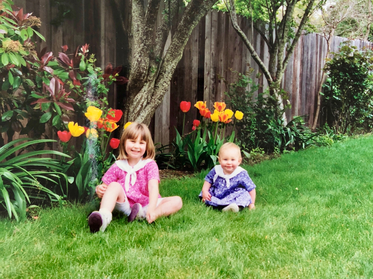 Two little girls wearing Easter dresses sitting on lawn in front of tulips.