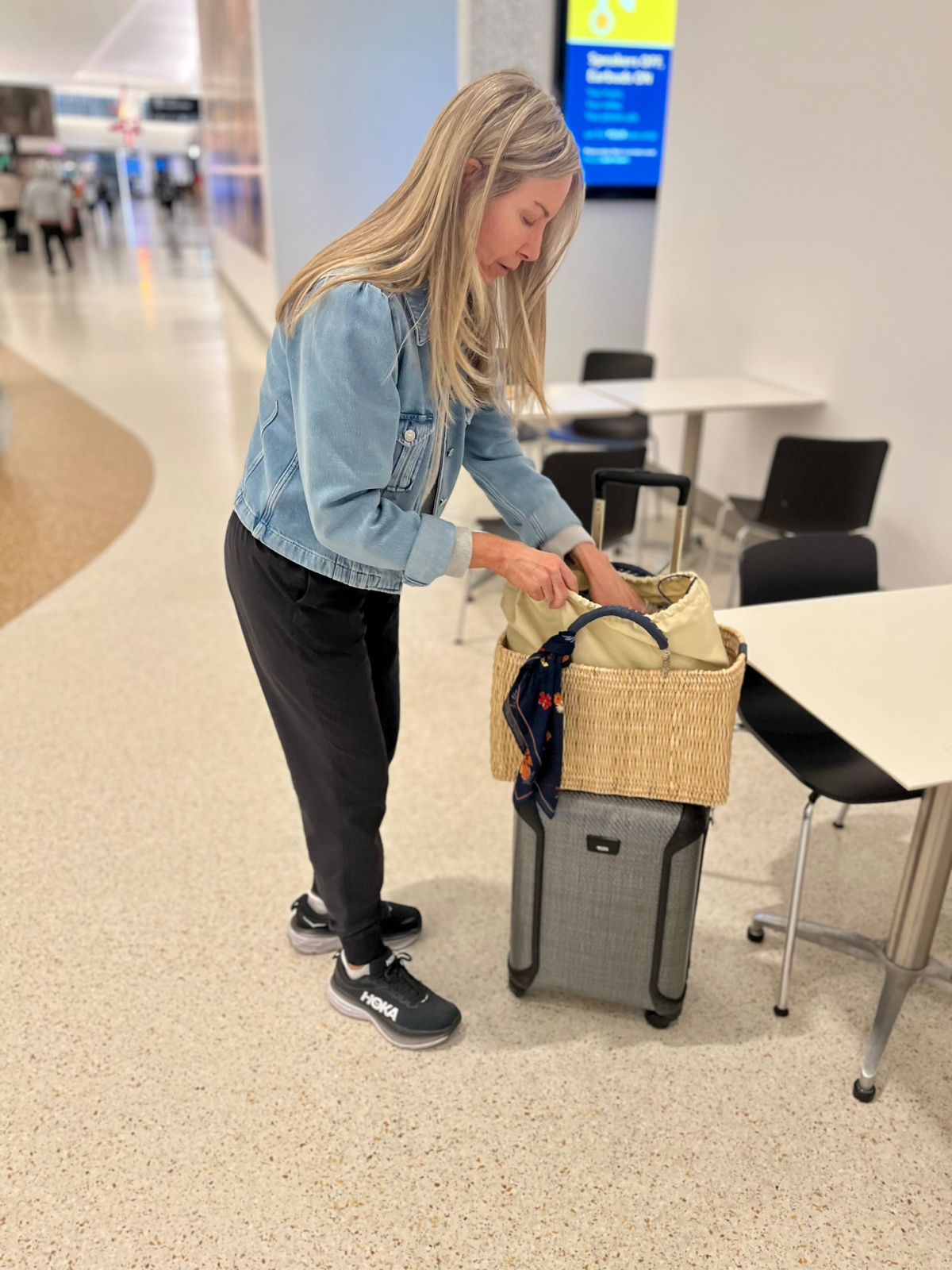 Woman leaning retrieving something from straw tote sitting on roller bag in airport.