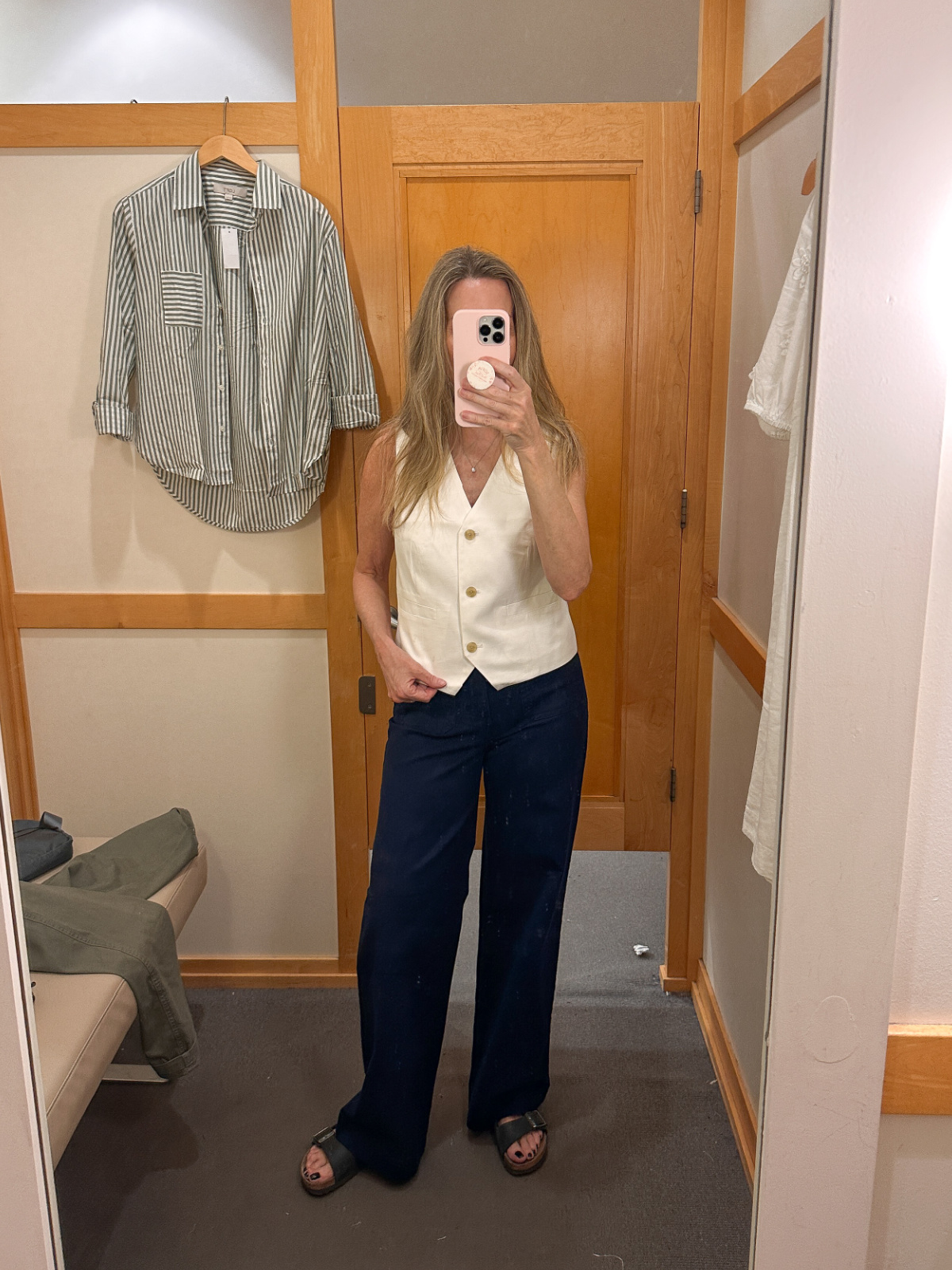 Woman taking dressing room mirror selfie wearing blue pants and white blouse.