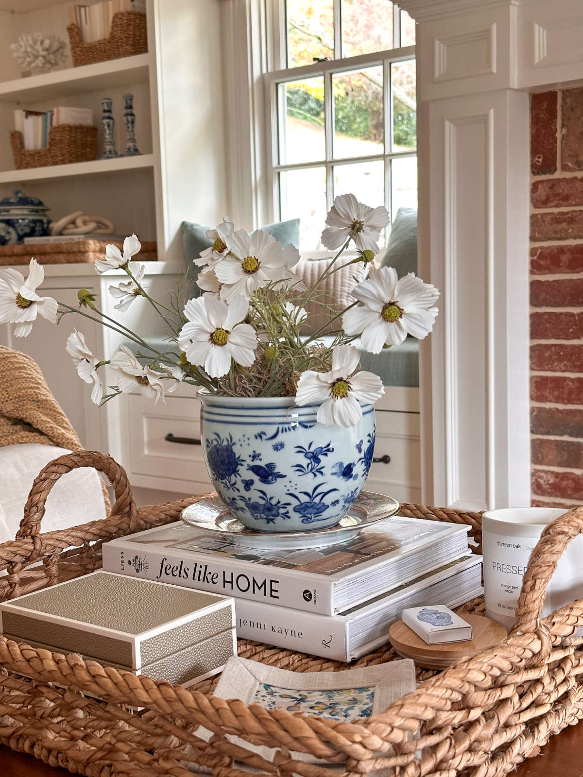 Square woven tray on coffee table filled with books, blue and white vase of white flowers, a rectangular box, candle and cocktail napkins.