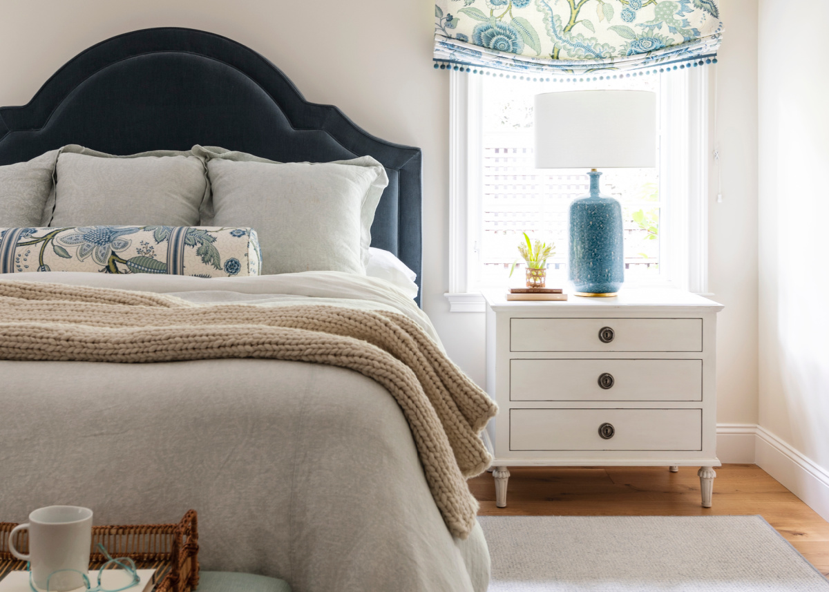 View into primary bedroom with blue velvet bed, linen bedding and ivory nightstand with blue table lamp.