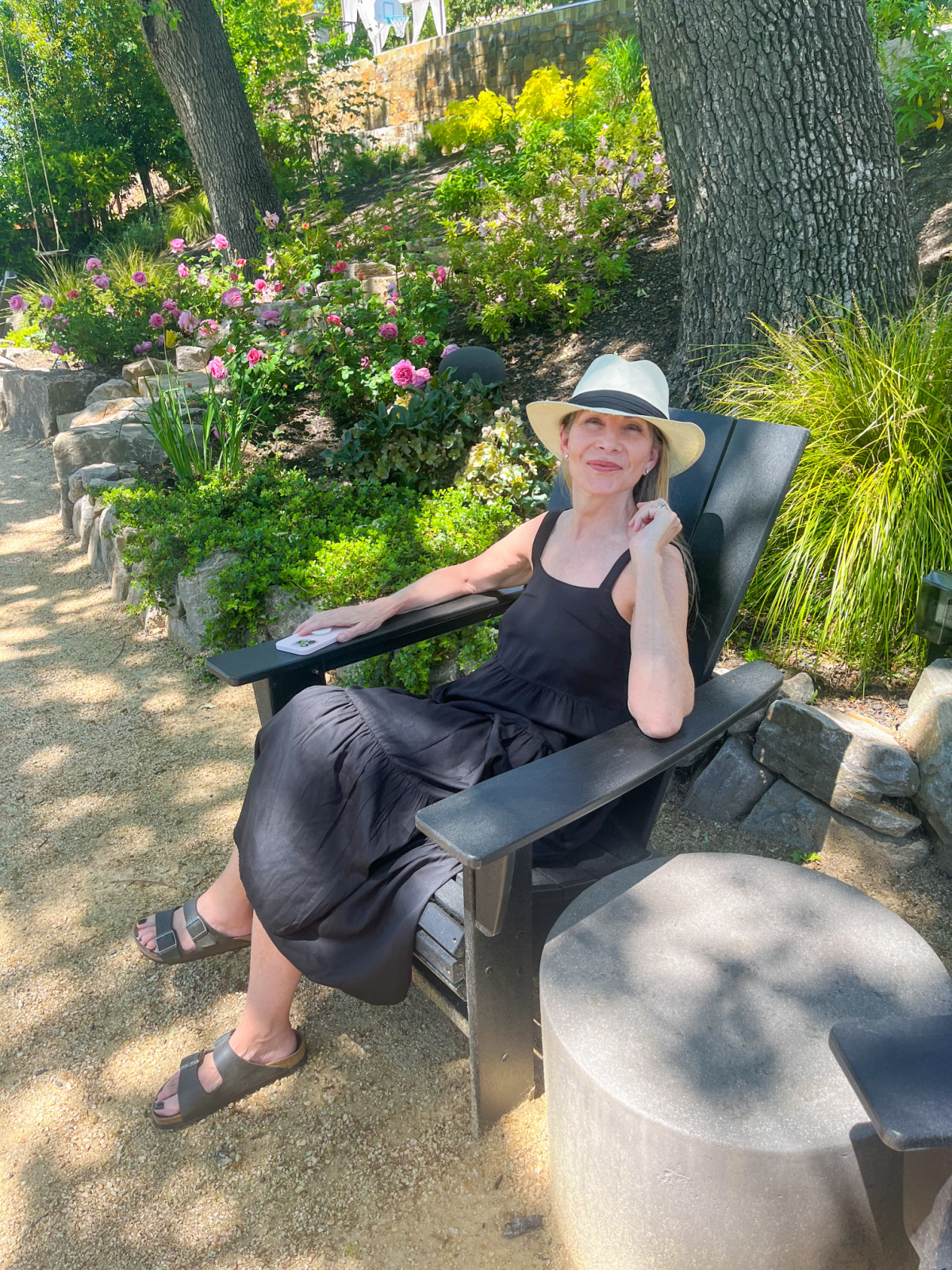 Woman wearing black dress and hat sitting in Adirondack chair in summer garden.