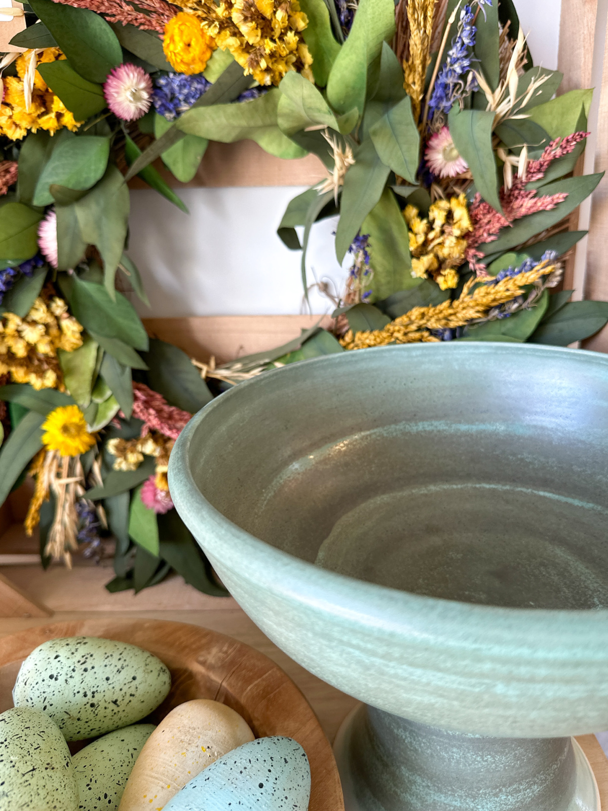 Pretty green bowl next to colorful eggs and a wildflower wreath.