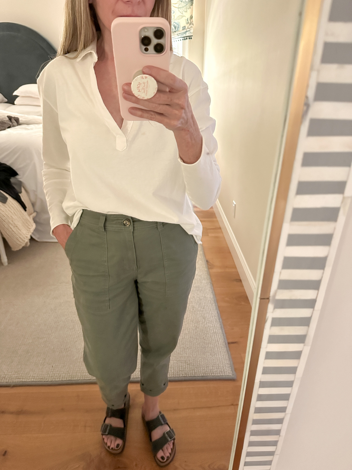 Woman wearing long sleeved white henley tee shirt and khaki green pants with Birkenstocks taking a mirror selfie.