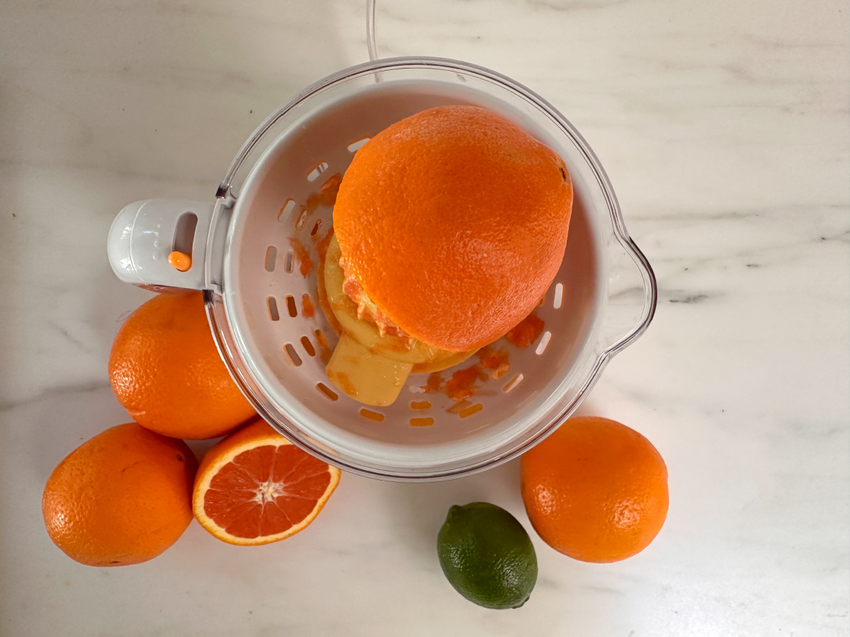 Overhead shot of orange in juicer with oranges and a lime surrounding it on kitchen counter.