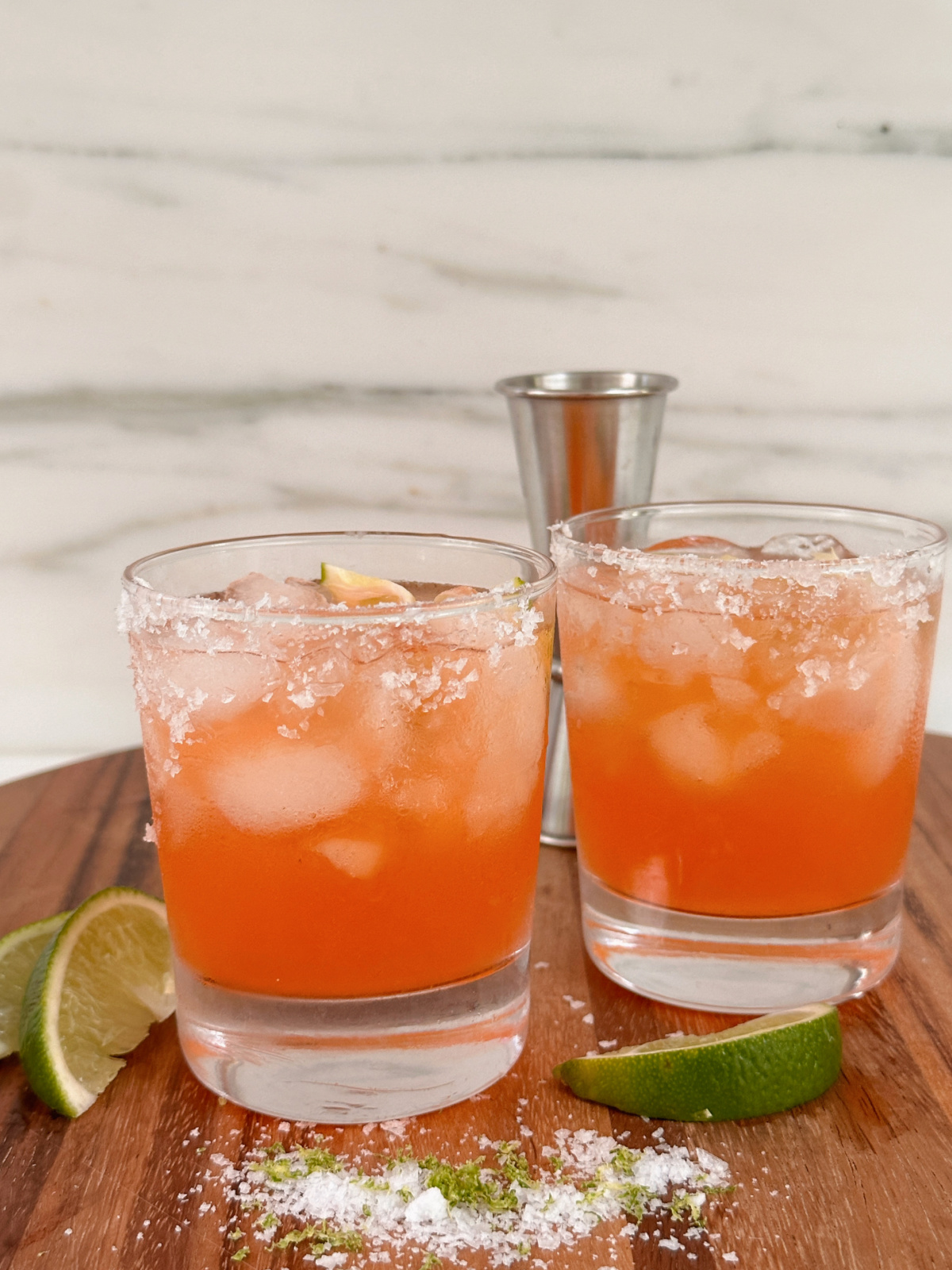 Two glasses of Aperol Margaritas on a wood board surrounded by lime wedges, flake salt and a jigger.