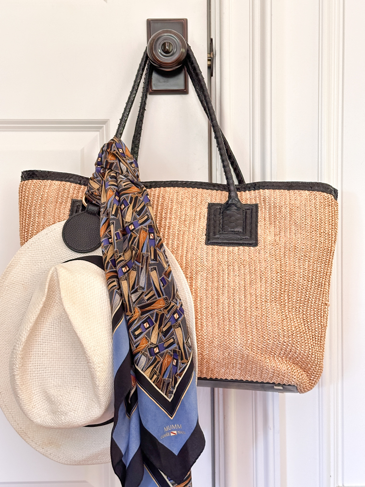 Straw tote with leather trim hanging on door knob along with hat and scarf.
