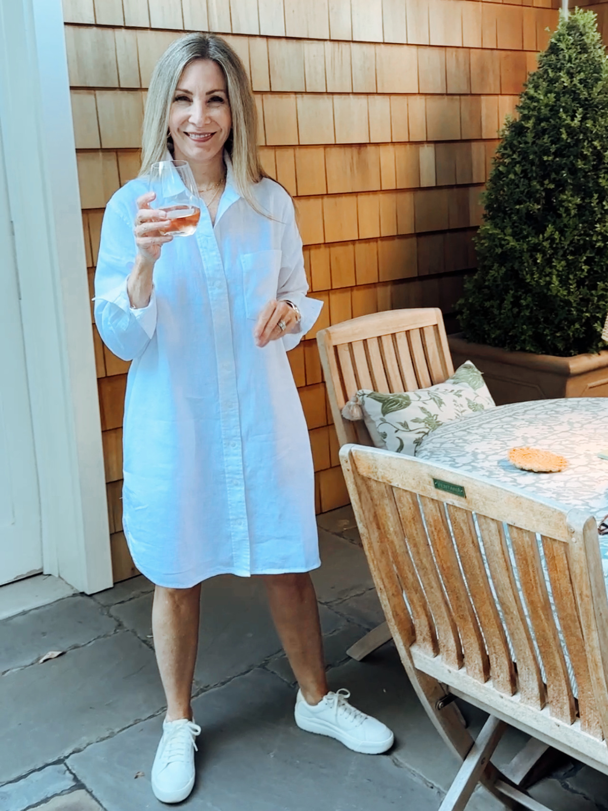 Woman wearing white shirt dress and sneakers standing on patio with glass of wine.